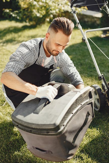 A man with neatly styled hair and a short beard is engaged in garden maintenance outdoors on a sunny day. He is wearing a checked shirt, black apron, and white gloves, and is kneeling on a well-maintained grassy lawn. In front of him is a black grass collection box attached to a lawn mower, which he appears to be emptying or inspecting. The background features lush green trees and a hedge, with sunlight filtering through the leaves, creating a bright and natural setting. Nearby, a lawn mower handlebar rests against the grass, indicating ongoing lawn care work. The scene exemplifies meticulous garden upkeep, including lawn mowing and grass collection, aligning with landscaping and hedge trimming tasks that Gardeners Chiswick might undertake, as referenced on the page titled 'Chiswick High Road garden maintenance guide for homeowners.'