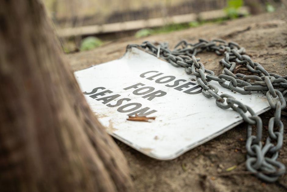A weathered white temporary sign lying on the ground in a garden, partially obscured by a large tree trunk in the foreground. The sign is chained down with thick metal links and reads 'Closed for Shason' in bold black letters, indicating restricted access. The surrounding area features moist, dark soil with patches of moss and scattered leaves, along with neatly maintained garden beds bordered by wooden planks. In the background, there are trimmed hedges and a pathway, suggesting a landscaped outdoor space undergoing routine maintenance or landscaping work. The lighting is natural, casting soft shadows across the scene, and reflecting a quiet, well-kept garden environment managed with professional care, such as that provided by Gardeners Chiswick.